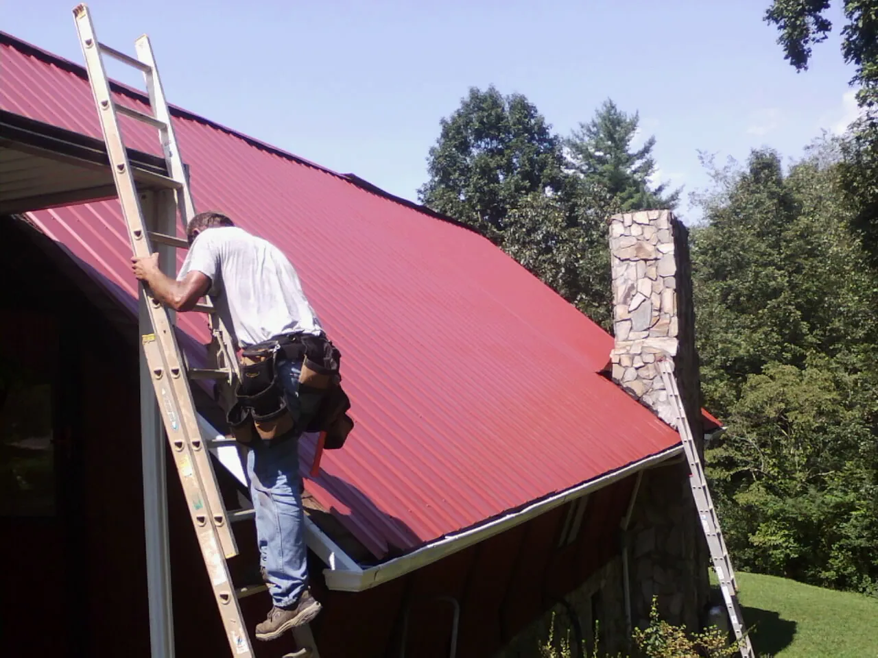Roofer on a ladder beside a red metal roof and stone chimney — Soffit & Fascia Repair by Roofing Brothers in Carrollton